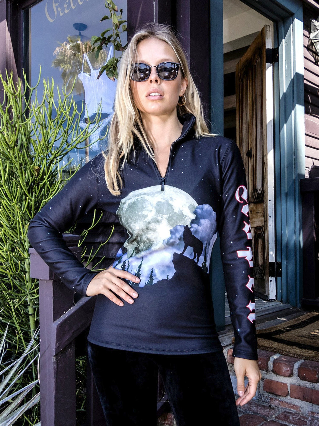 Woman in sunglasses wearing the Howling Wolf mid-layer top with a howling wolf under a full moon on black, standing on a porch.