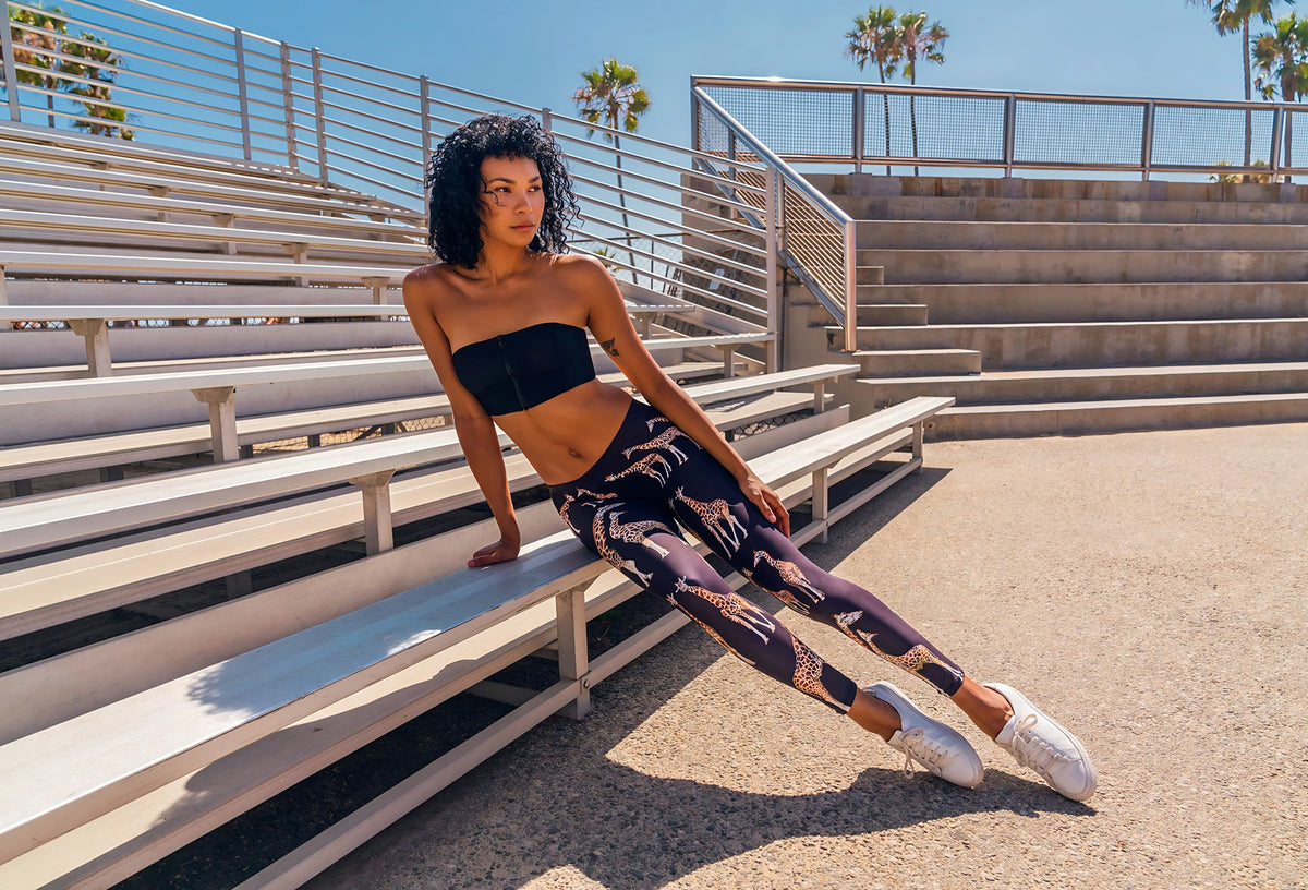 Woman sitting on outdoor bleachers wearing Giraffe Safari print leggings with giraffe artwork, black tube top, palm trees behind.
