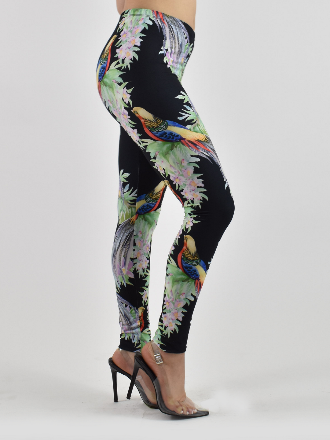 Side view of Denimaxx tropical bird print leggings with colorful pheasants and pink blossoms on black fabric, modeled with heels in a studio.