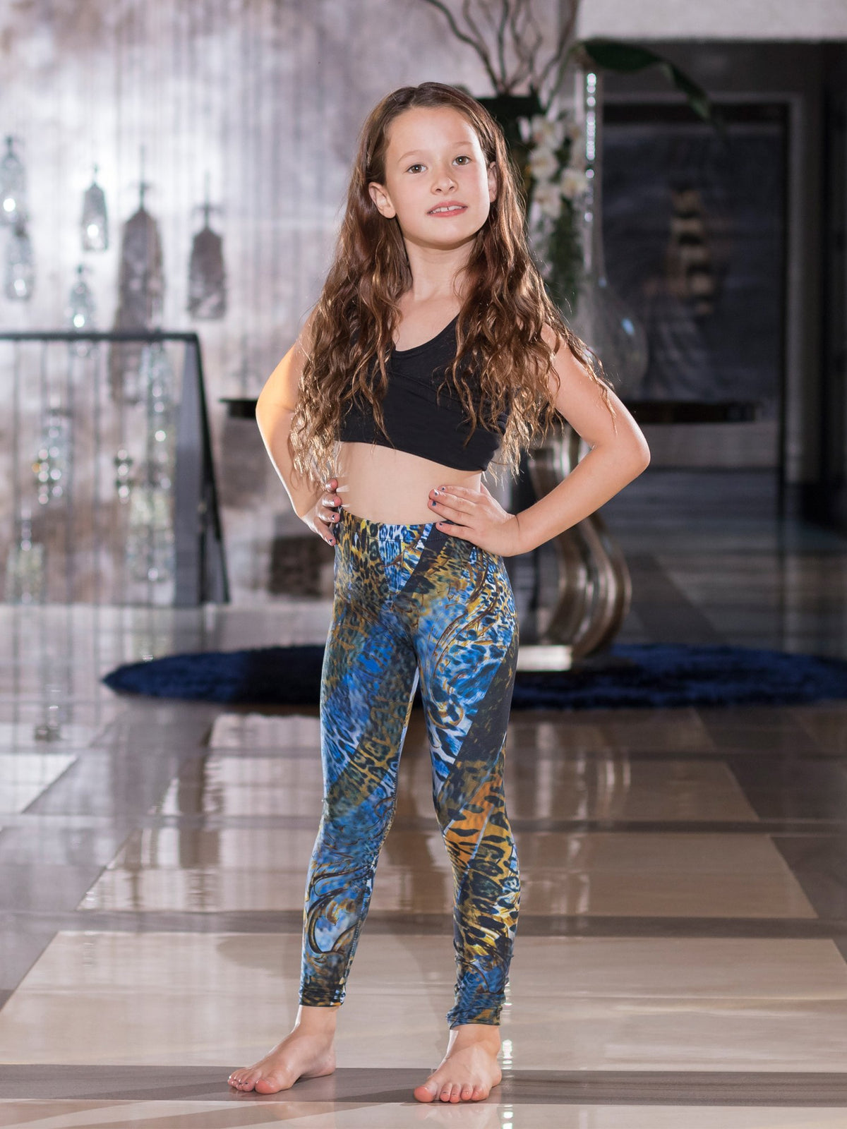Young girl with hands on hips wearing kids Blue Snake print leggings with blue and gold snake scales, black crop top, barefoot indoors.