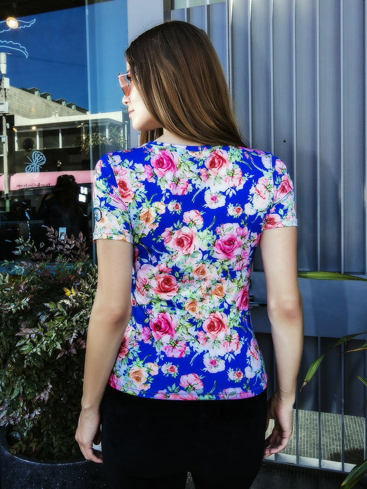 Back view of woman wearing the Denimaxx blue Roses short sleeve shirt with all-over pink rose print, outside a storefront.