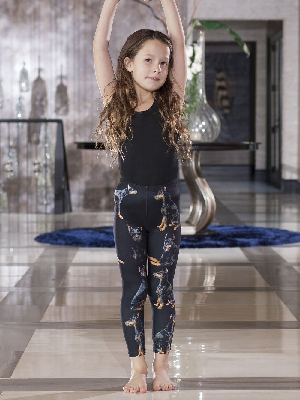 Young girl with arms raised overhead wearing Denimaxx kids Doberman Pinscher print leggings and a black tank top.