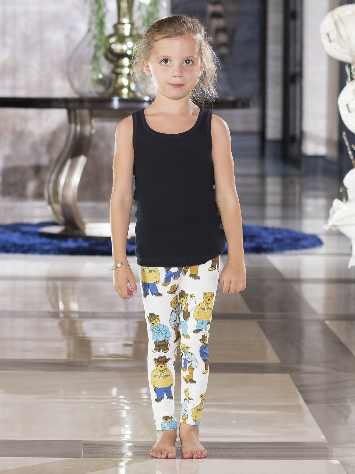 Young girl standing front-facing in kids Cowboy Teddy Bear print leggings with a black tank top indoors.