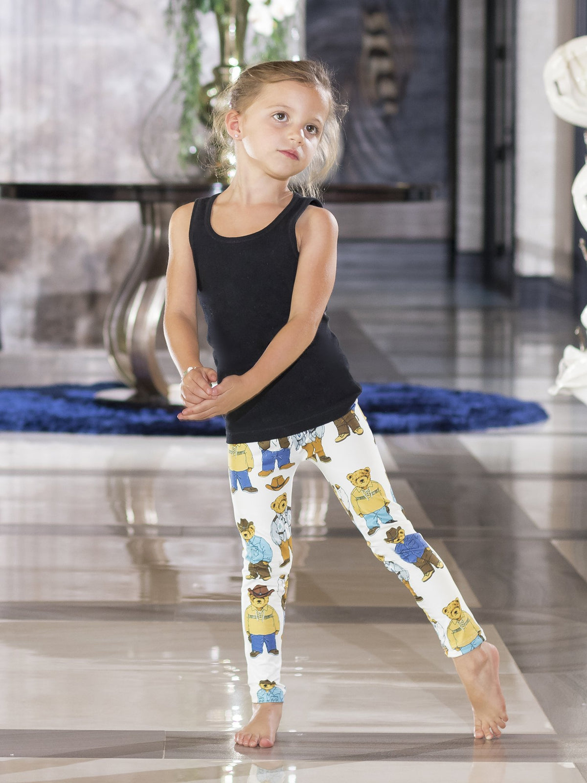 Young girl with one leg extended, modeling kids Cowboy Teddy Bear print leggings on a tiled floor.