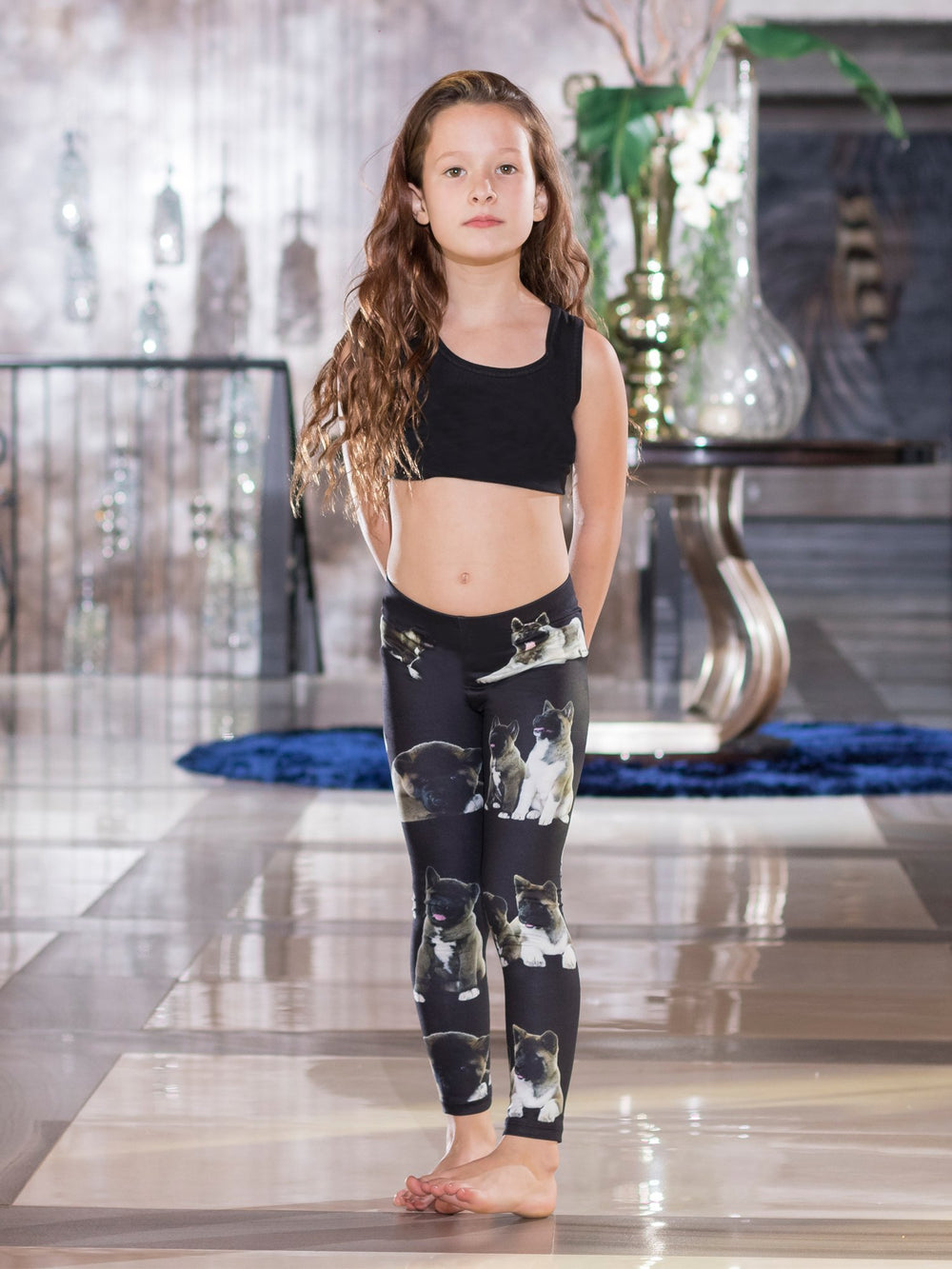 Young girl with hands behind her back wearing Denimaxx kids black Akita print graphic leggings and a black sports bra.