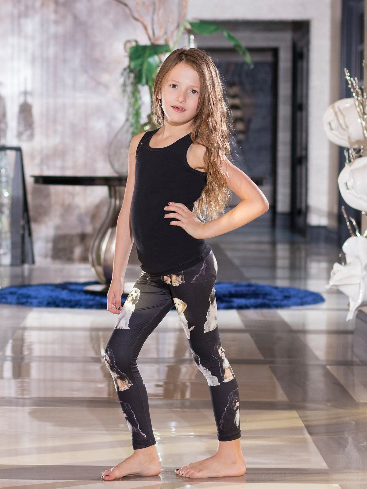 Young girl with hand on hip wearing kids Tibetan Terrier print leggings, white and black terrier faces on black, black tank top.