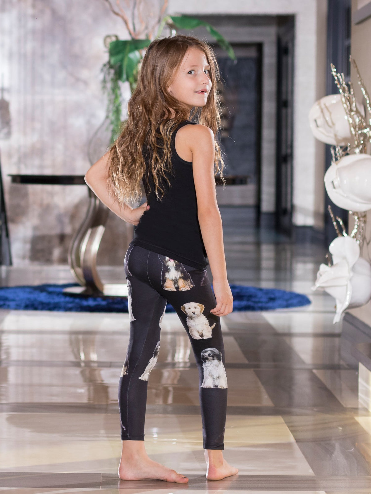 Side view of young girl with hand on hip looking back, wearing kids Tibetan Terrier print leggings, white terrier faces on black.
