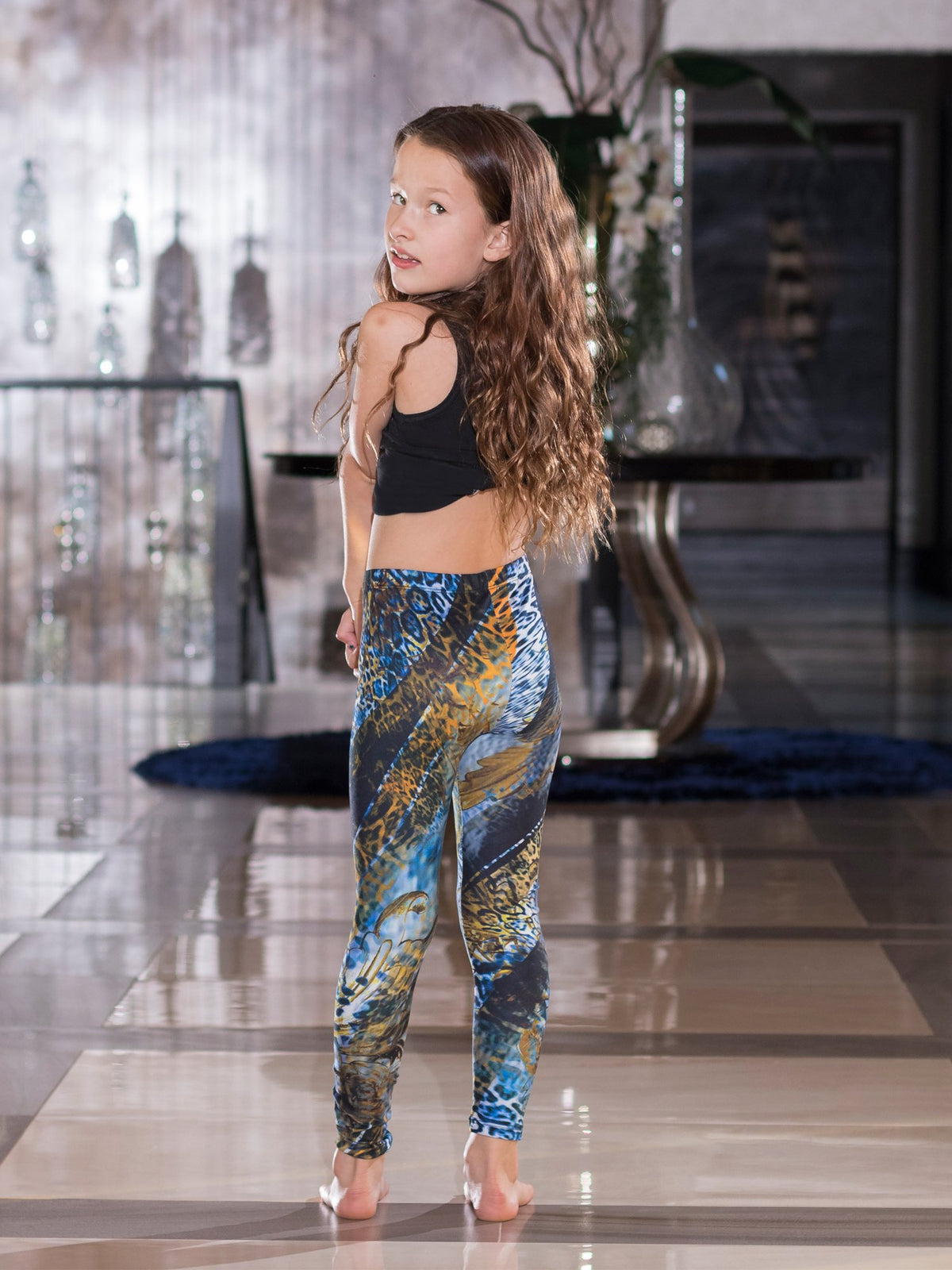 Young girl looking over her shoulder wearing kids Blue Snake print leggings with blue and gold snake scale print, back three-quarter view.