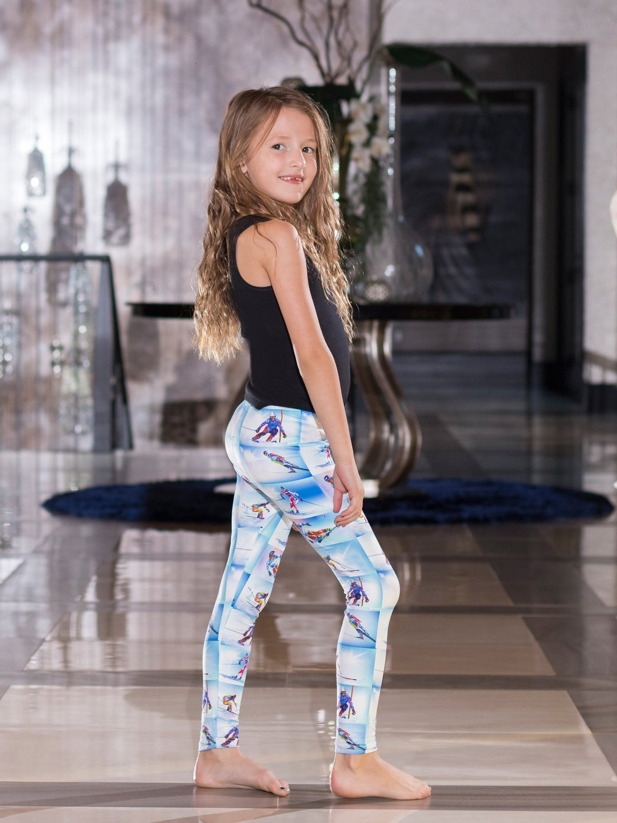 Back view of young girl looking over her shoulder, wearing kids Colorado Ski print leggings indoors.