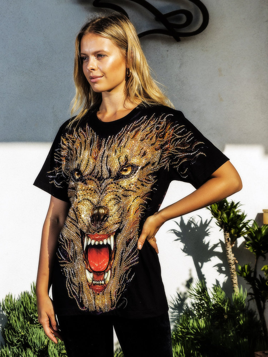Woman with hand on hip wearing the Denimaxx black Hell Hound beaded glow shirt with a beaded snarling hound face, plants beside a gray wall.