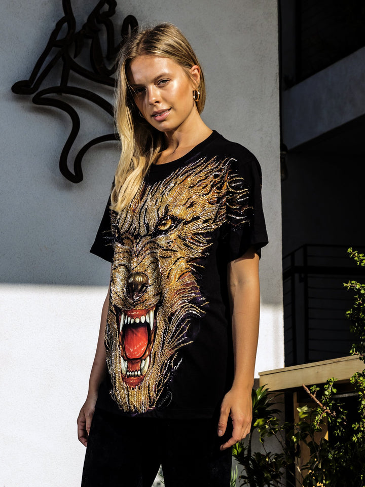 Woman wearing the Denimaxx black Hell Hound beaded glow shirt with a snarling beaded hound face, posed against a gray wall with metal art.