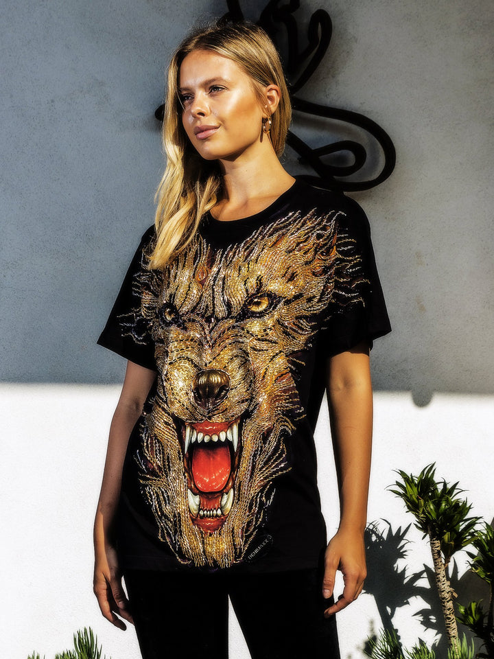 Woman modeling the Denimaxx black Hell Hound beaded glow shirt with gold and red beaded hound artwork, three-quarter view against a gray wall.
