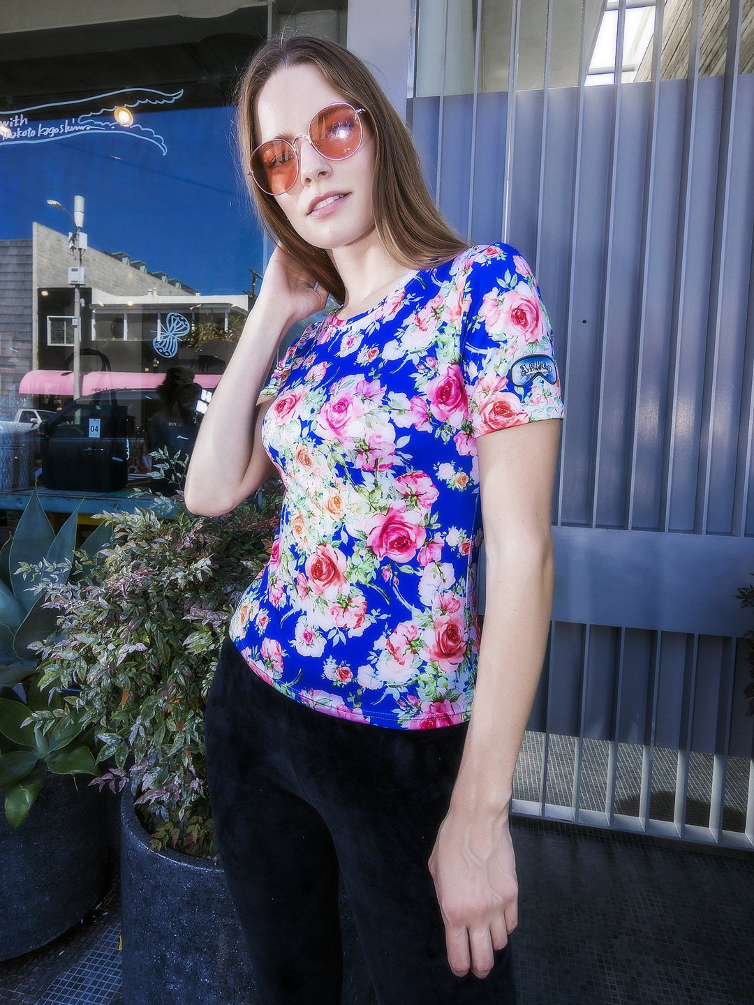 Woman with hand in her hair wearing the Denimaxx blue Roses short sleeve shirt with pink rose artwork, storefront backdrop.